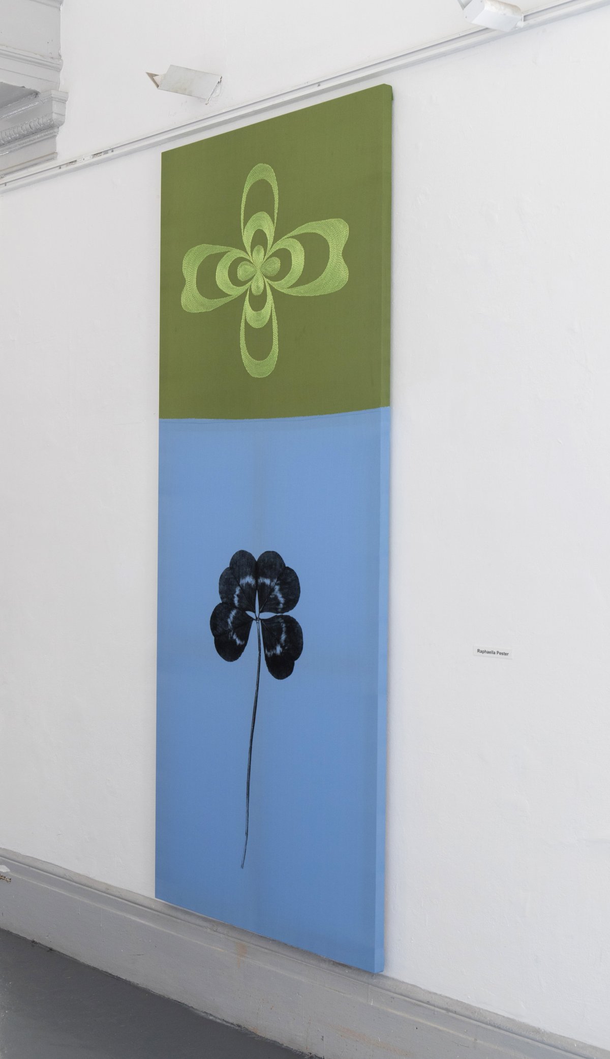 Installation photo of long rectangular painting, split in two: on the top, a light green floral motif on a darker green background; on the bottom a blue leaf/flower on a light blue background