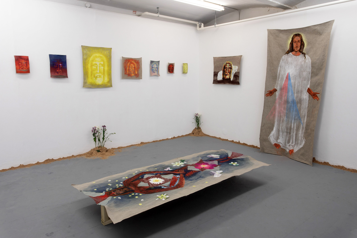 Space with religious paintings, including one like a shroud on the floor. Where the wall meets the floor there is sand and a couple of small mounds have flowers coming out of them.