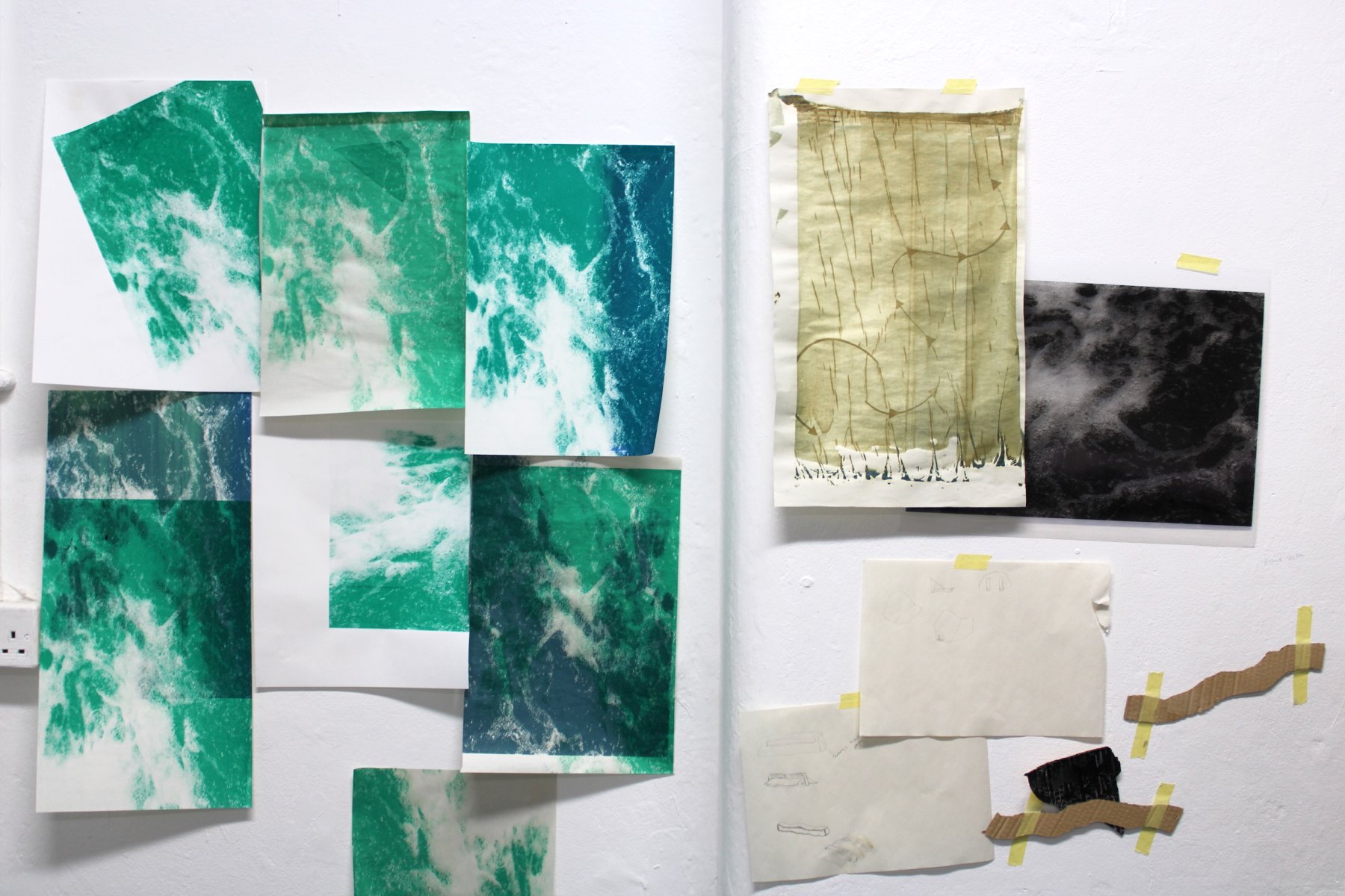 Image of wall with blue and green screenprint of water overlaid across the left side of the wall. On the right-hand side of the wall is one screenprint depicting arrows moving across the paper from bottom left to top right. On the right of the screenprint is a black and white image of water. Below are abstract cardboard forms and additional drawings on paper, all taped to the wall.