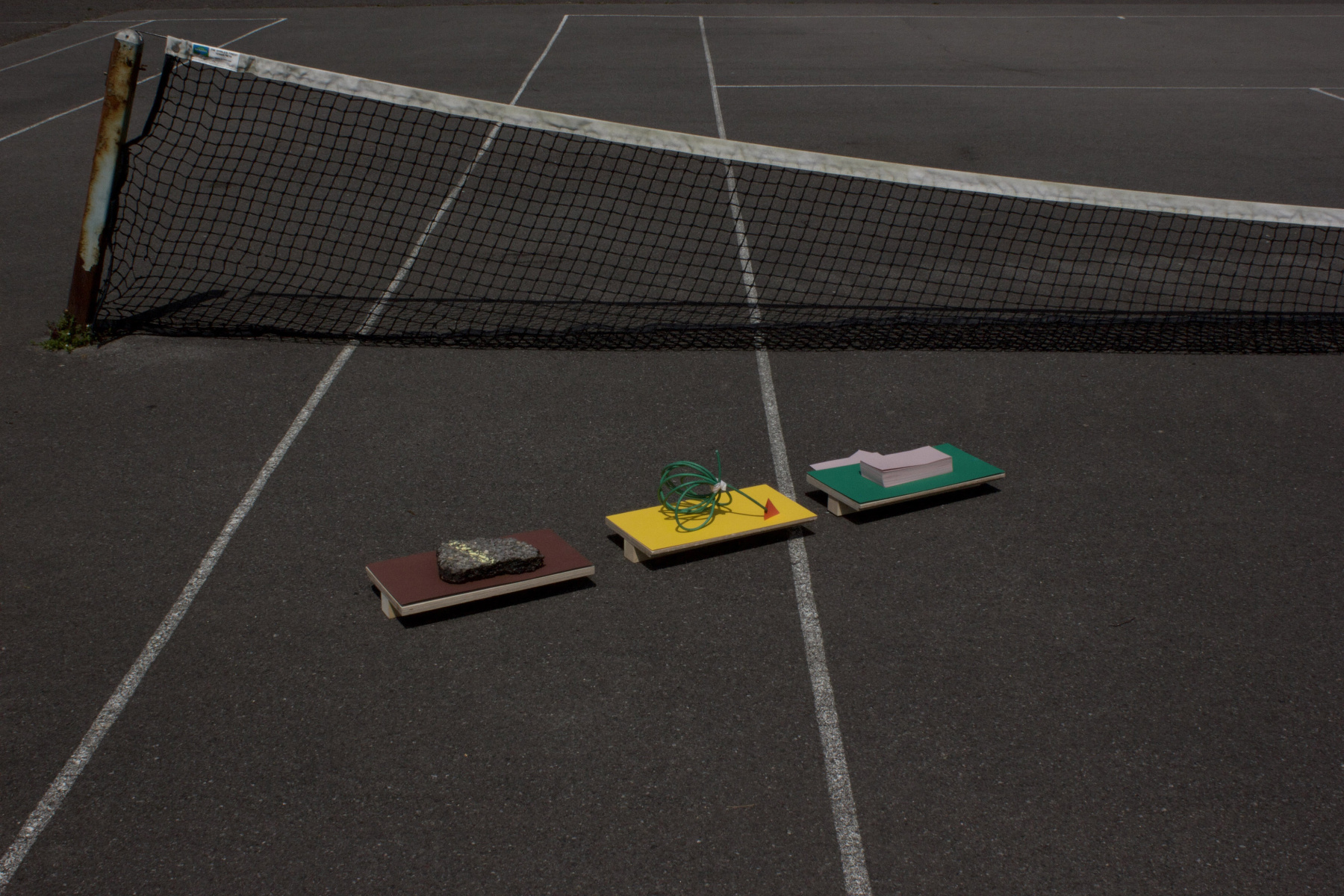 Three small platforms in brown, yellow and green, with objects on placed on the top, piece of tarmac, green wire and stack of pink paper, photographed in a tennis court.